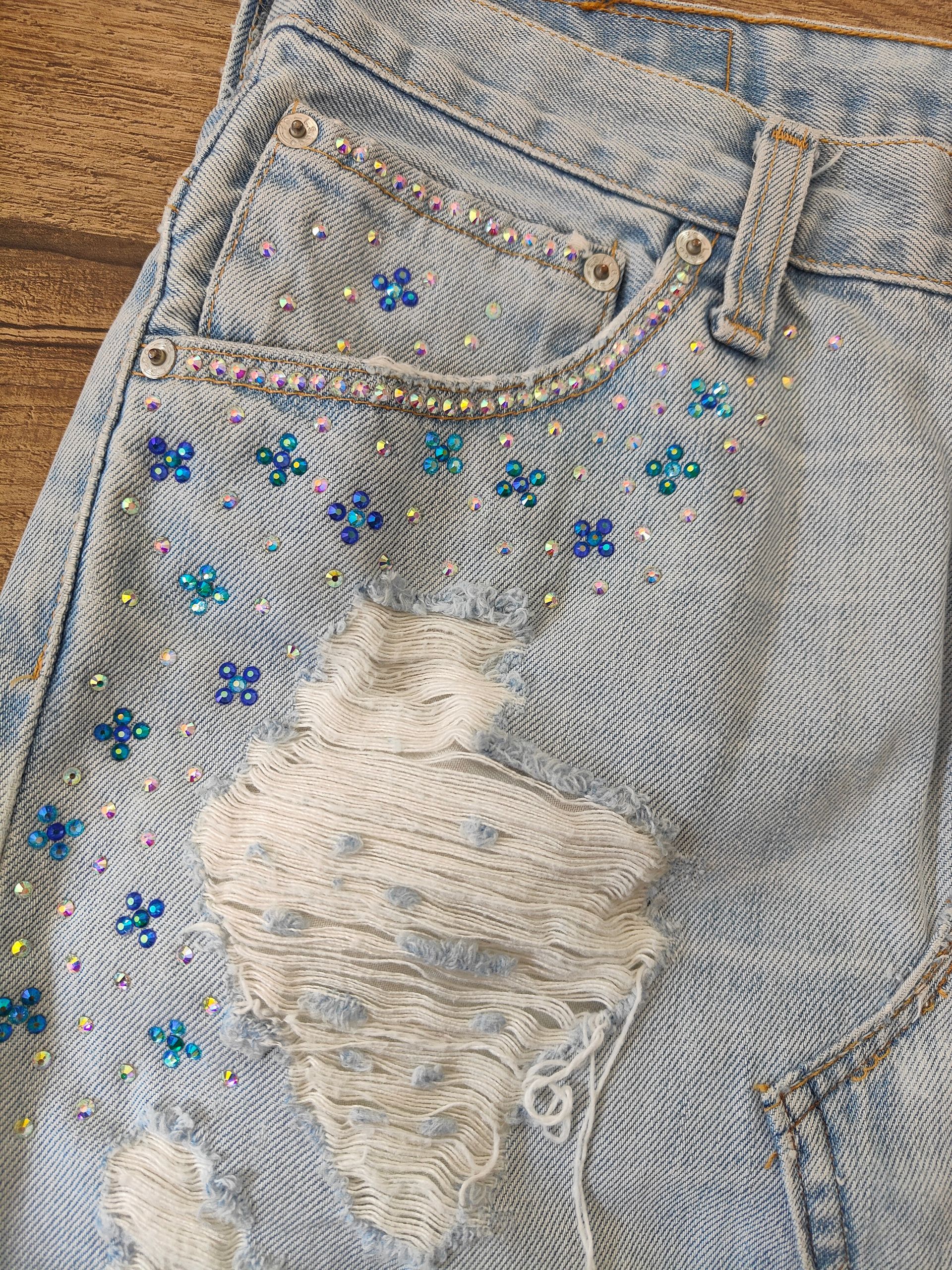 Jeans chiari effetto acid wash con strappi e scoloriture, decorati con piccole perline a forma di fiore blu e strass sparsi.