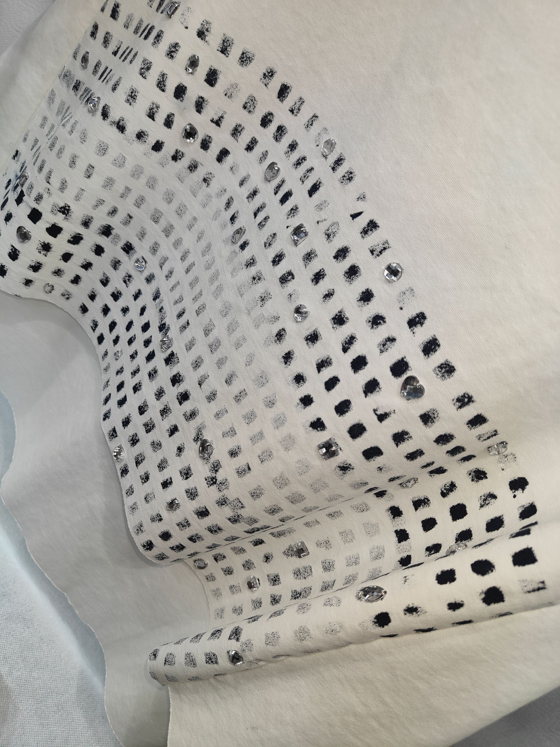 Primo piano di un tessuto color crema con un motivo di pois quadrati scuri dipinti a mano e piccoli strass sparsi.