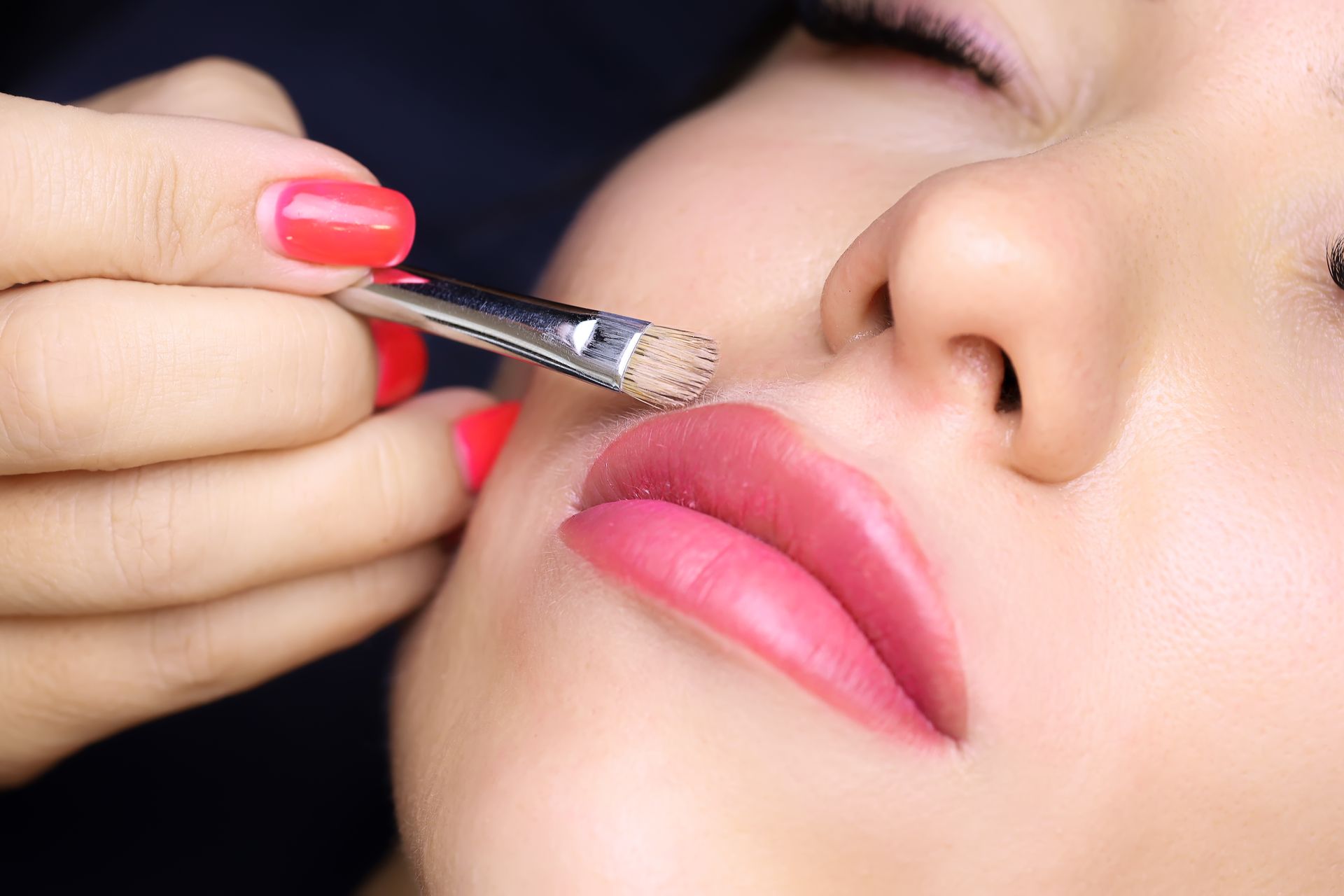 A woman with red nails is getting her lips painted with a brush.