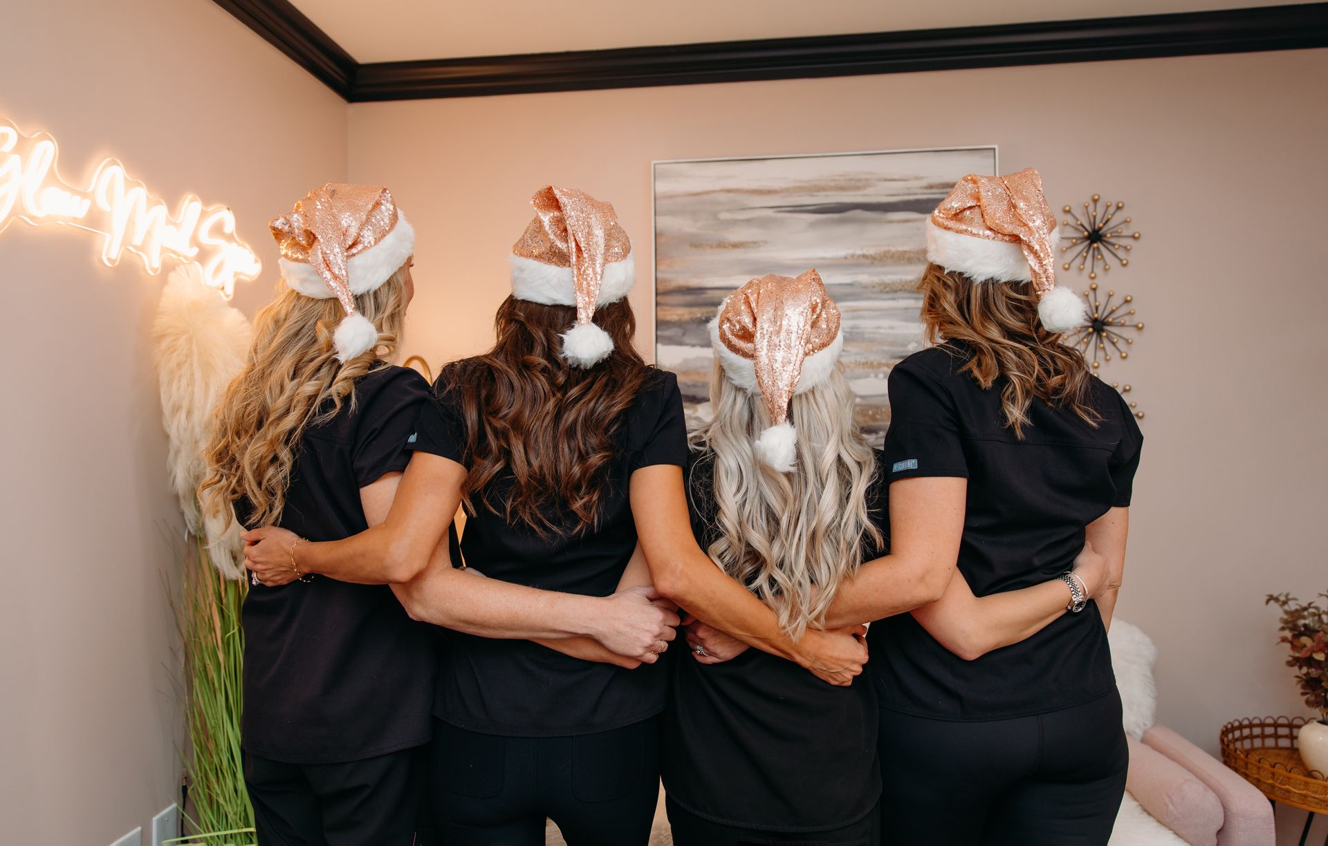 Four women in Santa hats arm-in-arm, facing away. They're in a room with decor, all wearing black shirts.