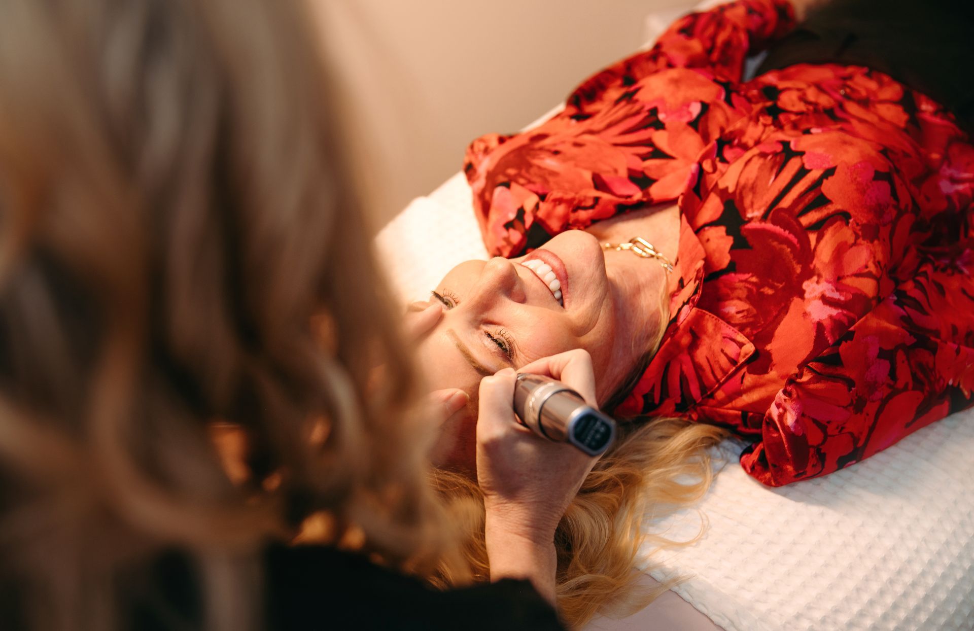 A woman is laying on a bed getting her eyebrows done by a makeup artist.