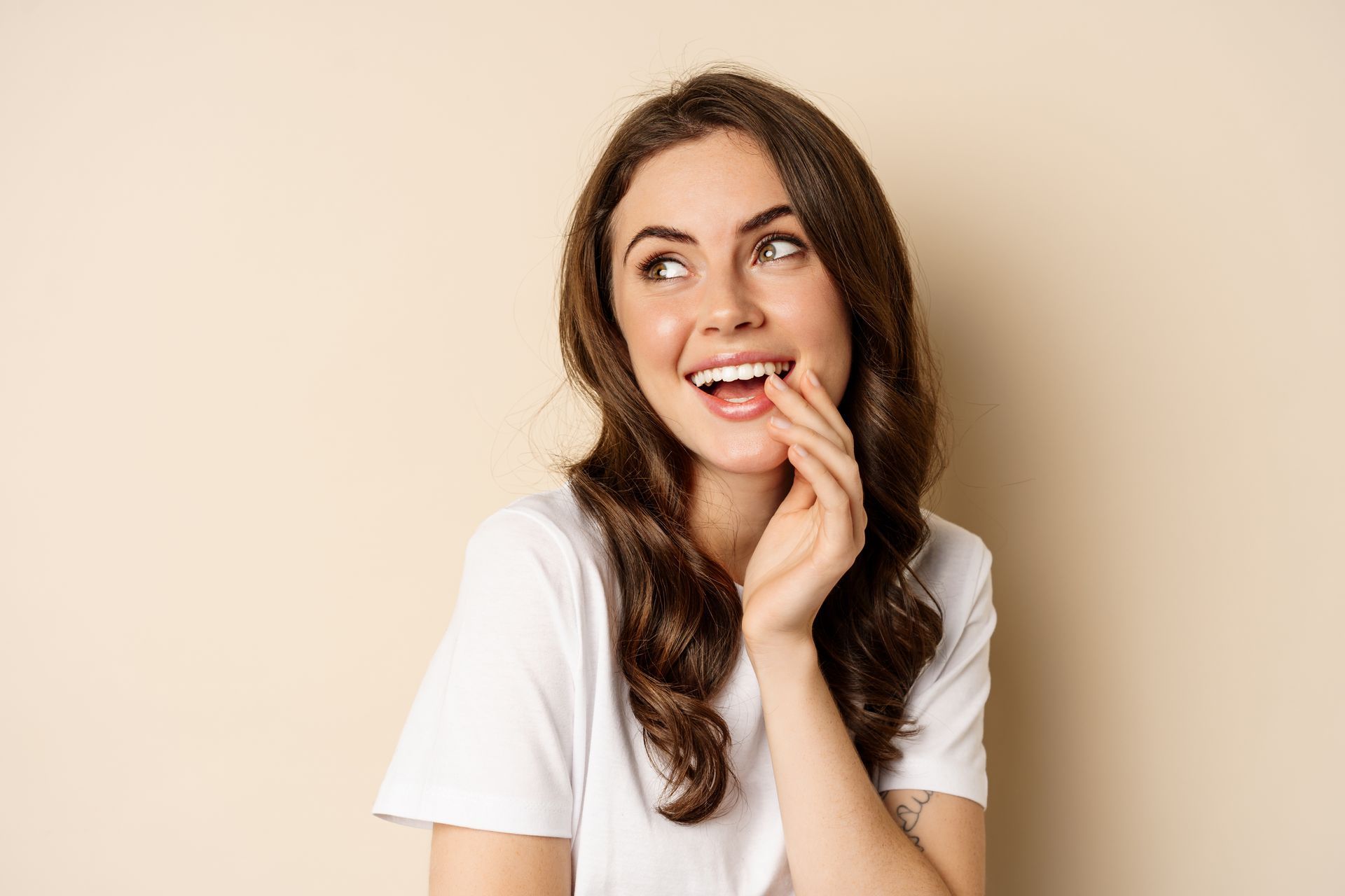 A woman in a white t-shirt is smiling and covering her mouth with her hand.