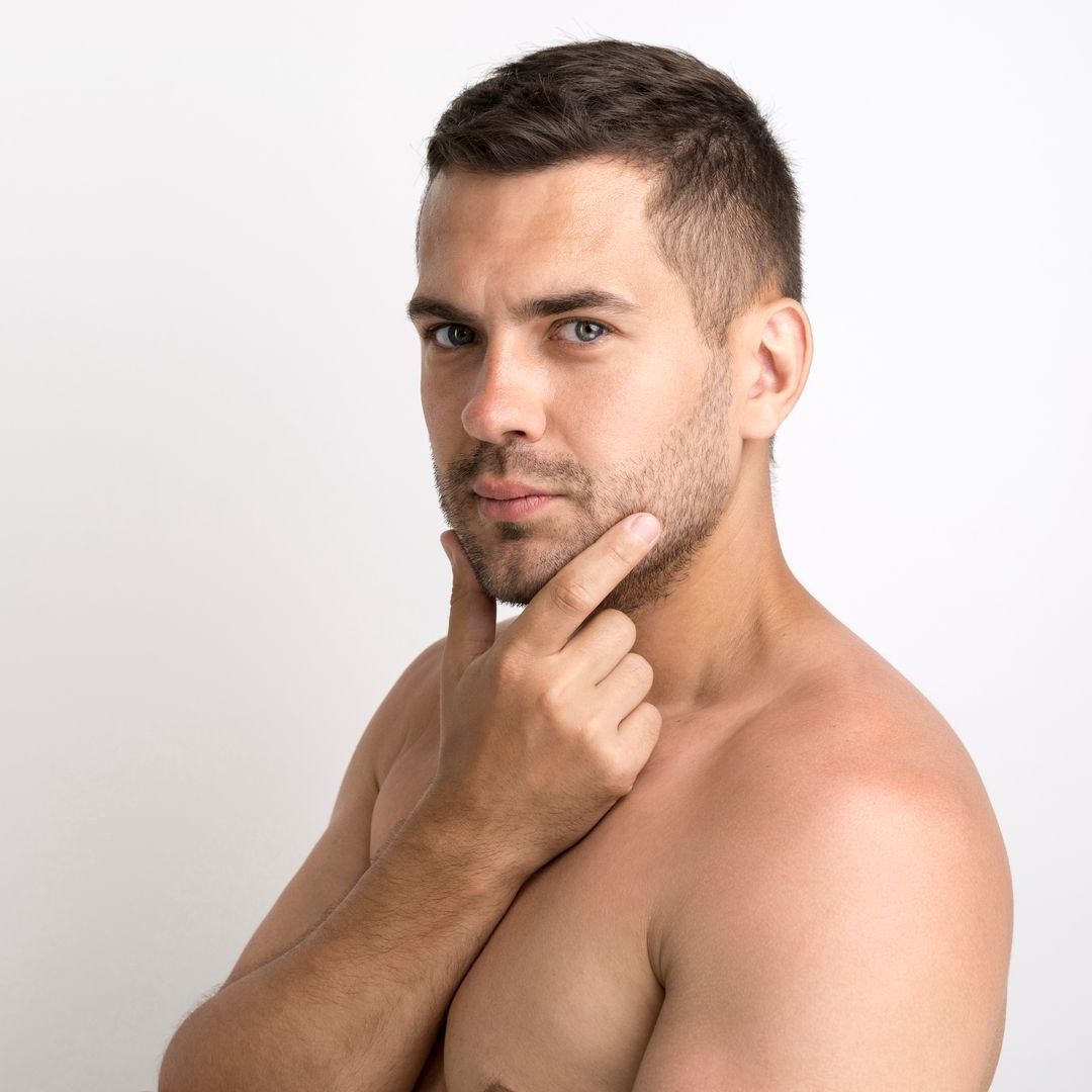 A shirtless man with a beard is holding his hand to his chin.