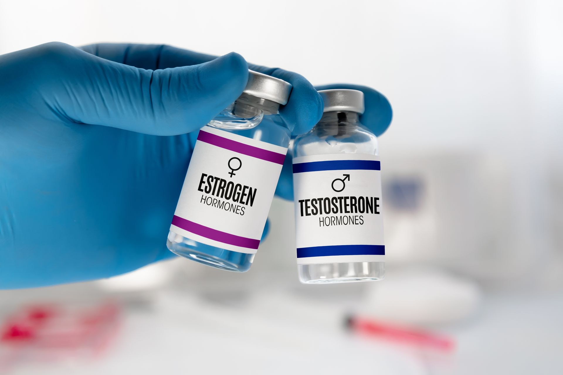 A hand in a blue glove is holding two bottles of testosterone and estrogen.