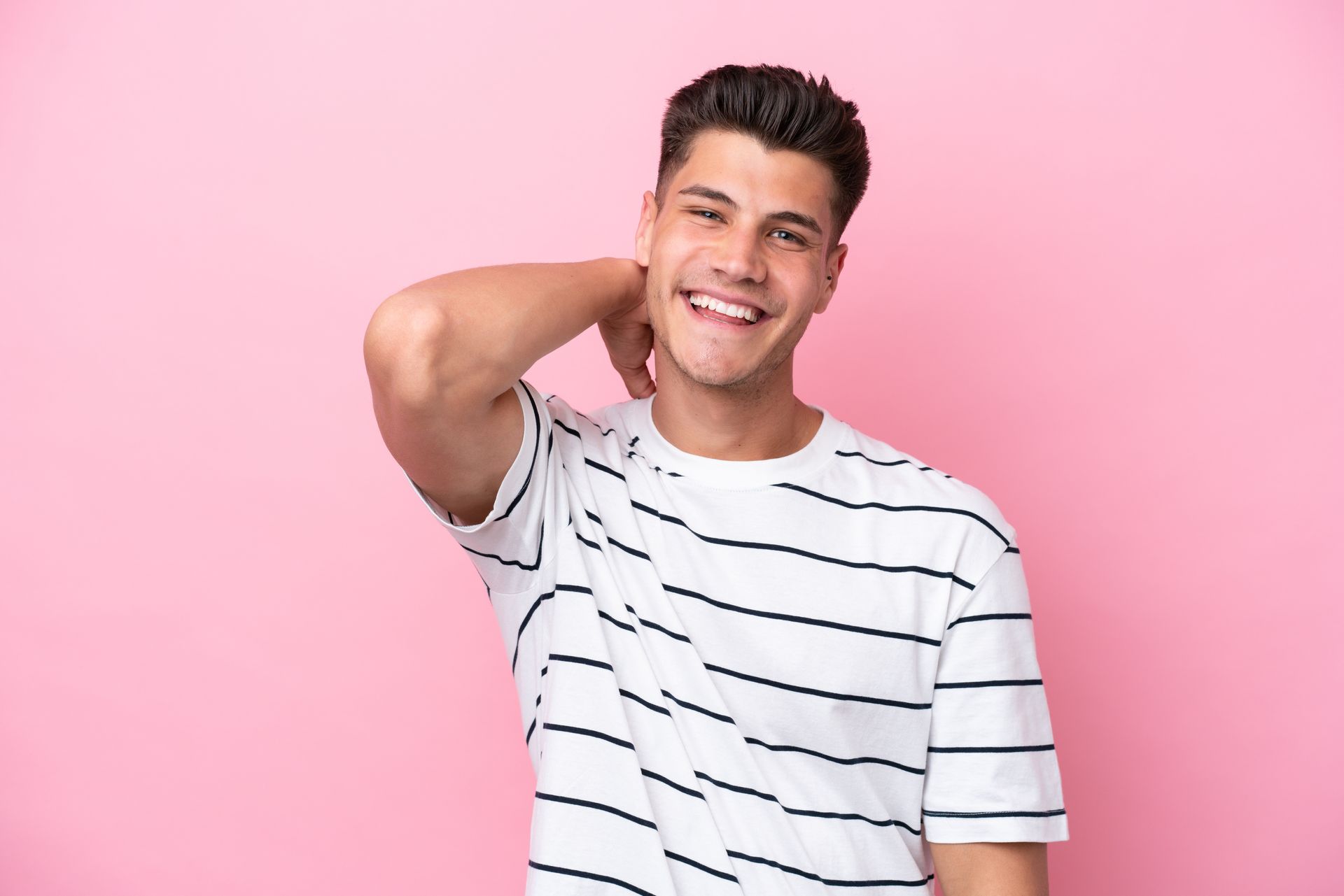A young man is smiling and touching his neck against a pink background.