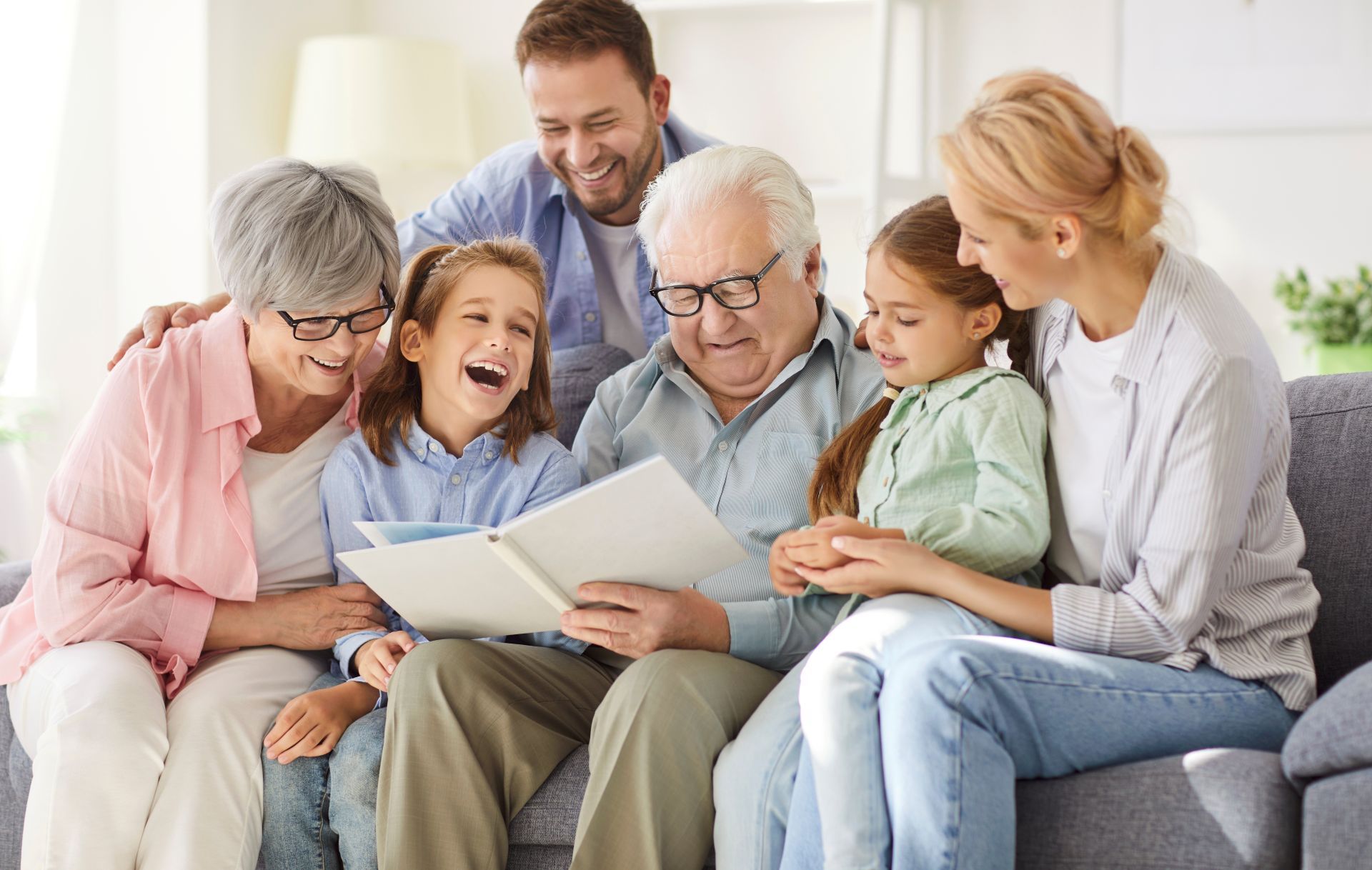 Family of seven smiles and looks at photo album on sofa indoors.