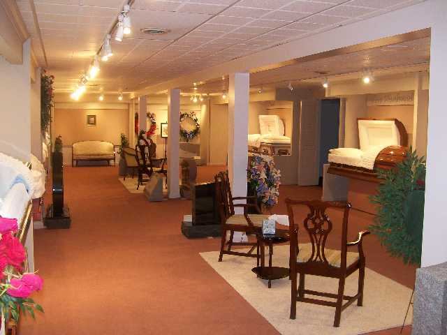 Funeral home showroom with caskets, seating, flowers, and displays. Beige carpet, white walls, and recessed lighting.