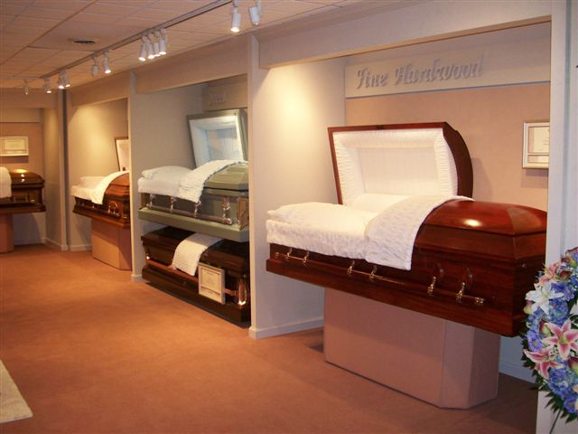 Inside a funeral home, several caskets of different colors are on display, with a floral arrangement in the corner.