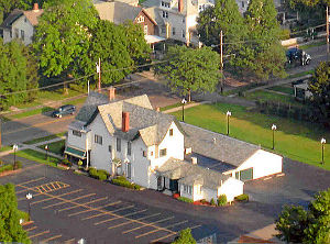 Aerial view of a two-story white building with a parking lot in front. Houses and trees surround it.