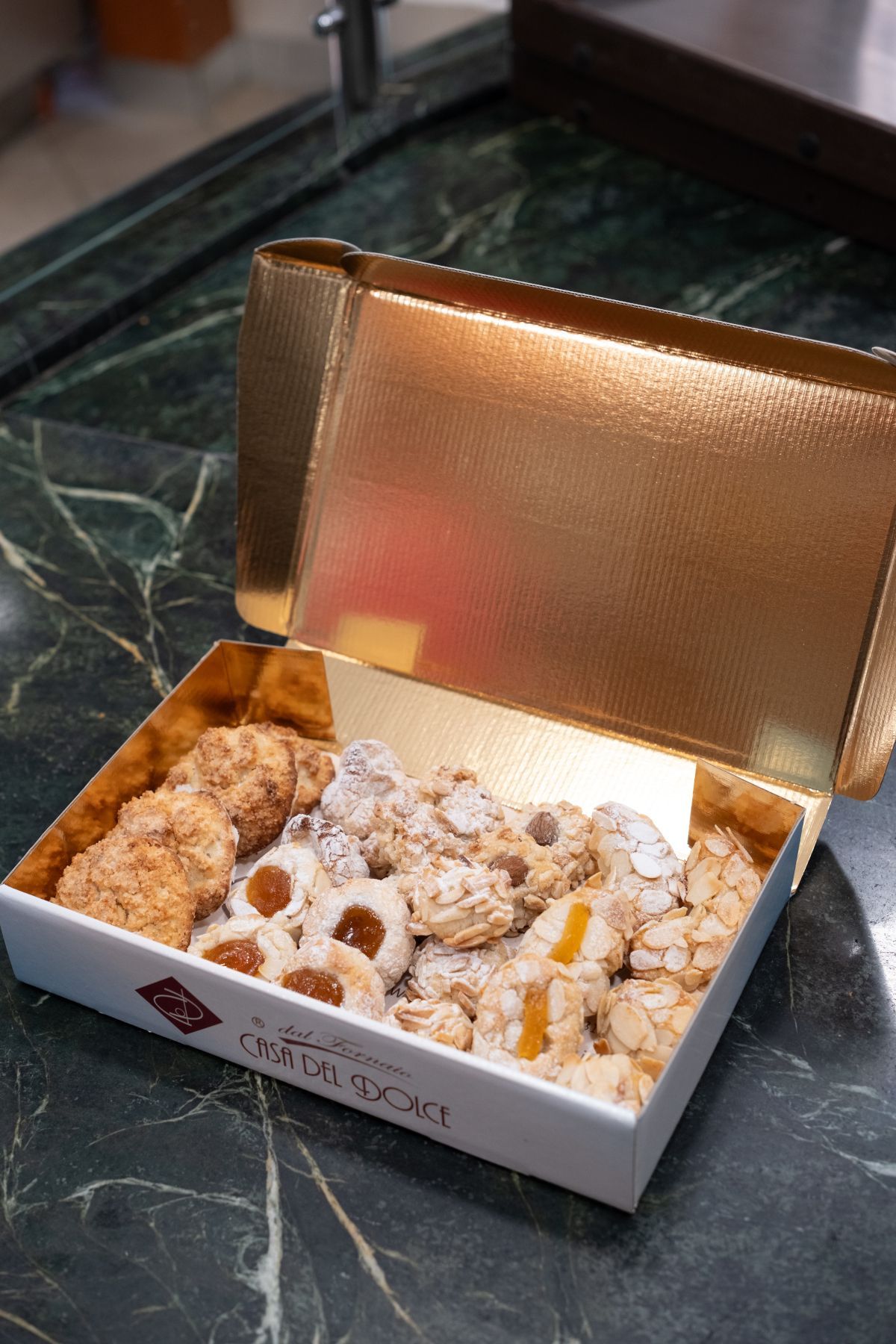 Scatola di biscotti assortiti con coperchio dorato, su una superficie di marmo verde.