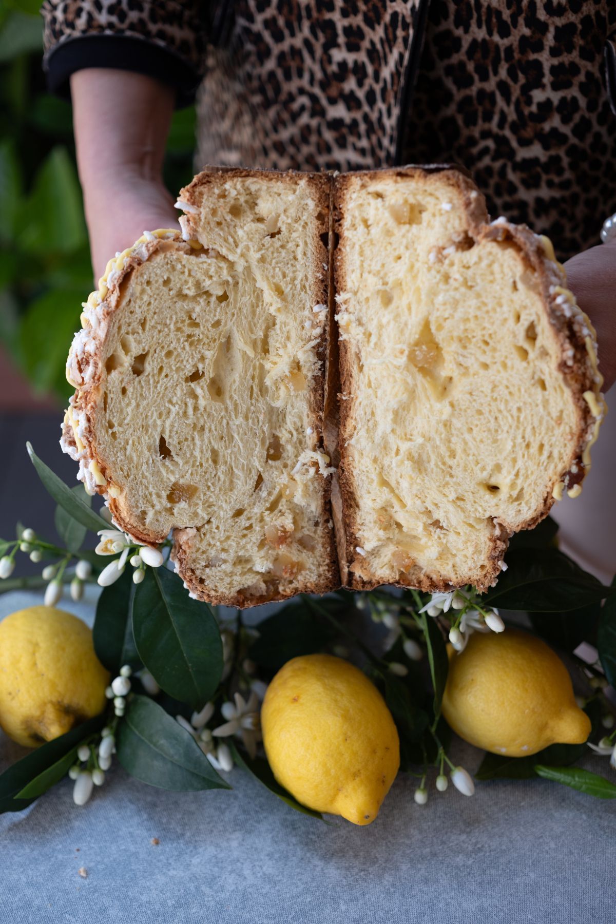 Persona che tiene in mano una torta brioche tagliata. Limoni e fiori la circondano su un tavolo.