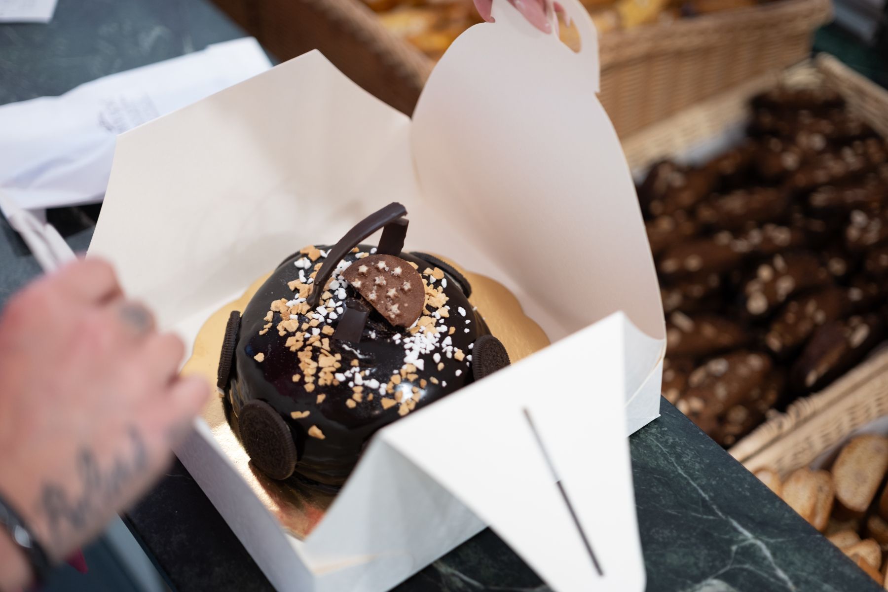 Torta al cioccolato in una scatola bianca, tenuta in mano, con altri prodotti da forno sullo sfondo.