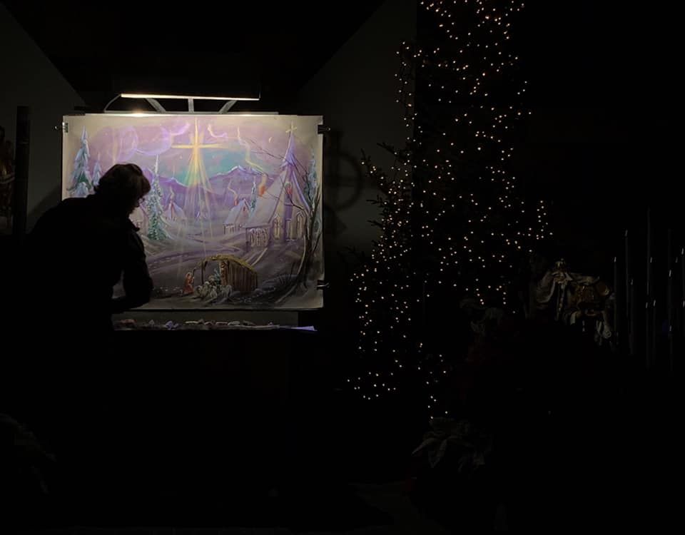 A woman is painting a picture in a dark room with a Christmas tree in the background.