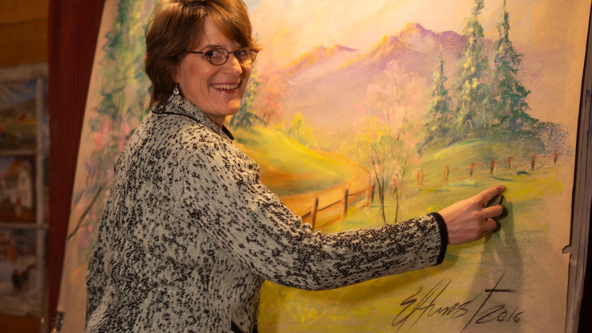 A woman is standing in front of a painting of a landscape.