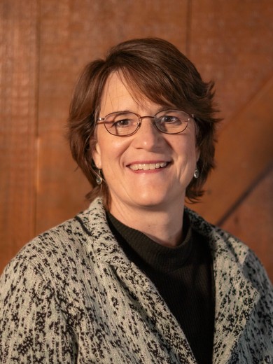 A woman wearing glasses and a jacket is smiling in front of a wooden wall.