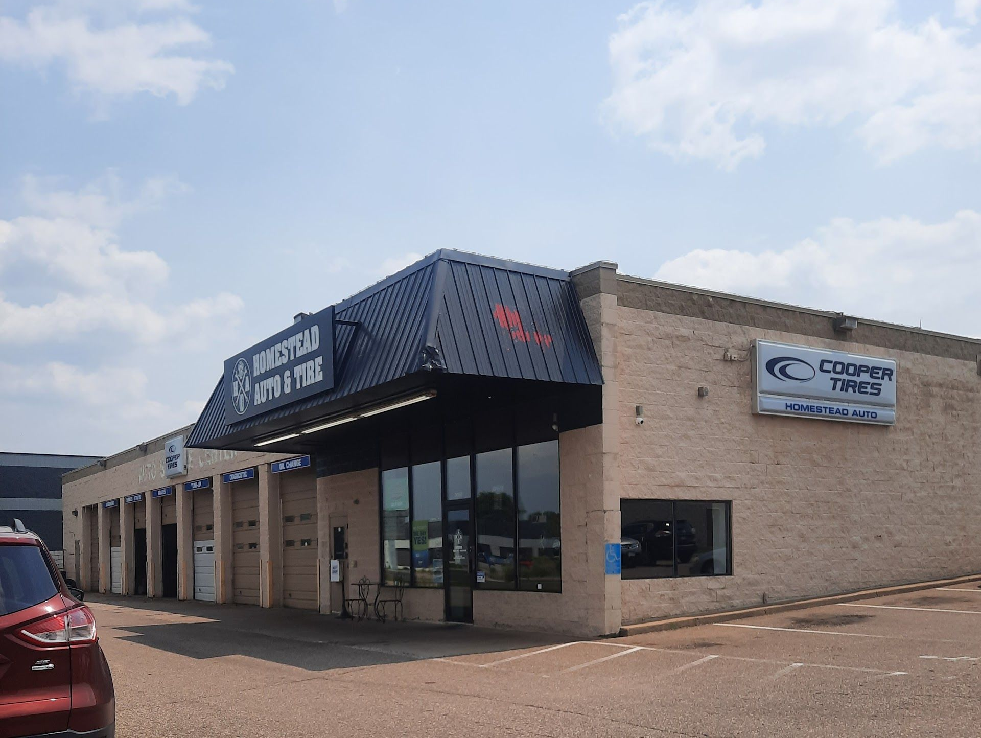 A street-level view of a tan brick automotive shop with a dark metal awning and a Cooper Tires sign on the side. | Homestead Auto & Tire