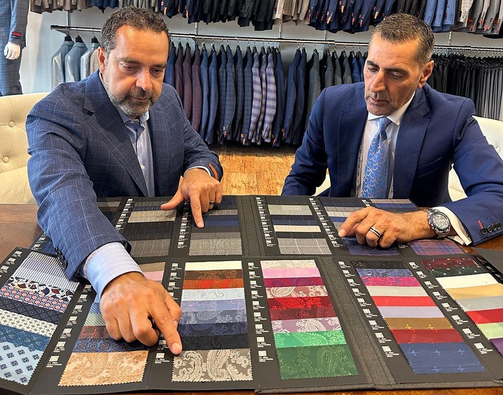 Two men in suits examining tie fabric swatches at a table in a suit shop.