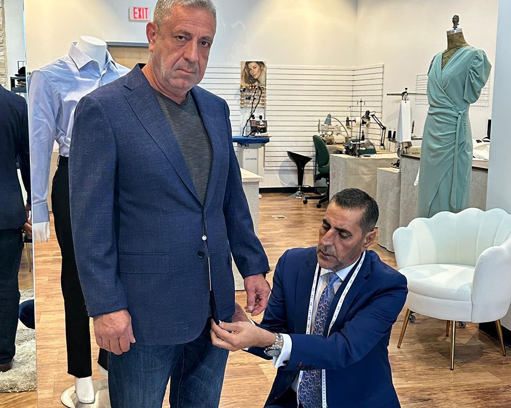 Tailor measuring a man's suit in a shop. The man wears a blazer, jeans, and a grey shirt.