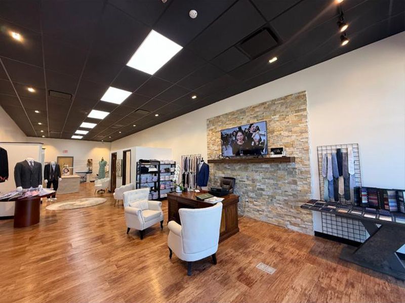 Interior of a men's clothing store: wooden floor, stone wall with TV, suits on display, white chairs, and ties.