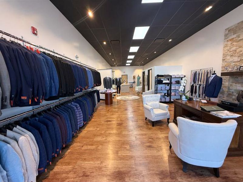 Inside a menswear store, suits in various blues hang on racks; white armchairs and a desk are visible.