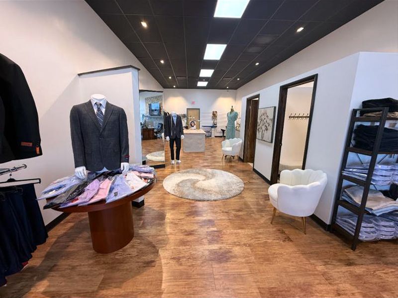 Men's clothing store interior with mannequins wearing suits and shirts, wood floor, and round rug.