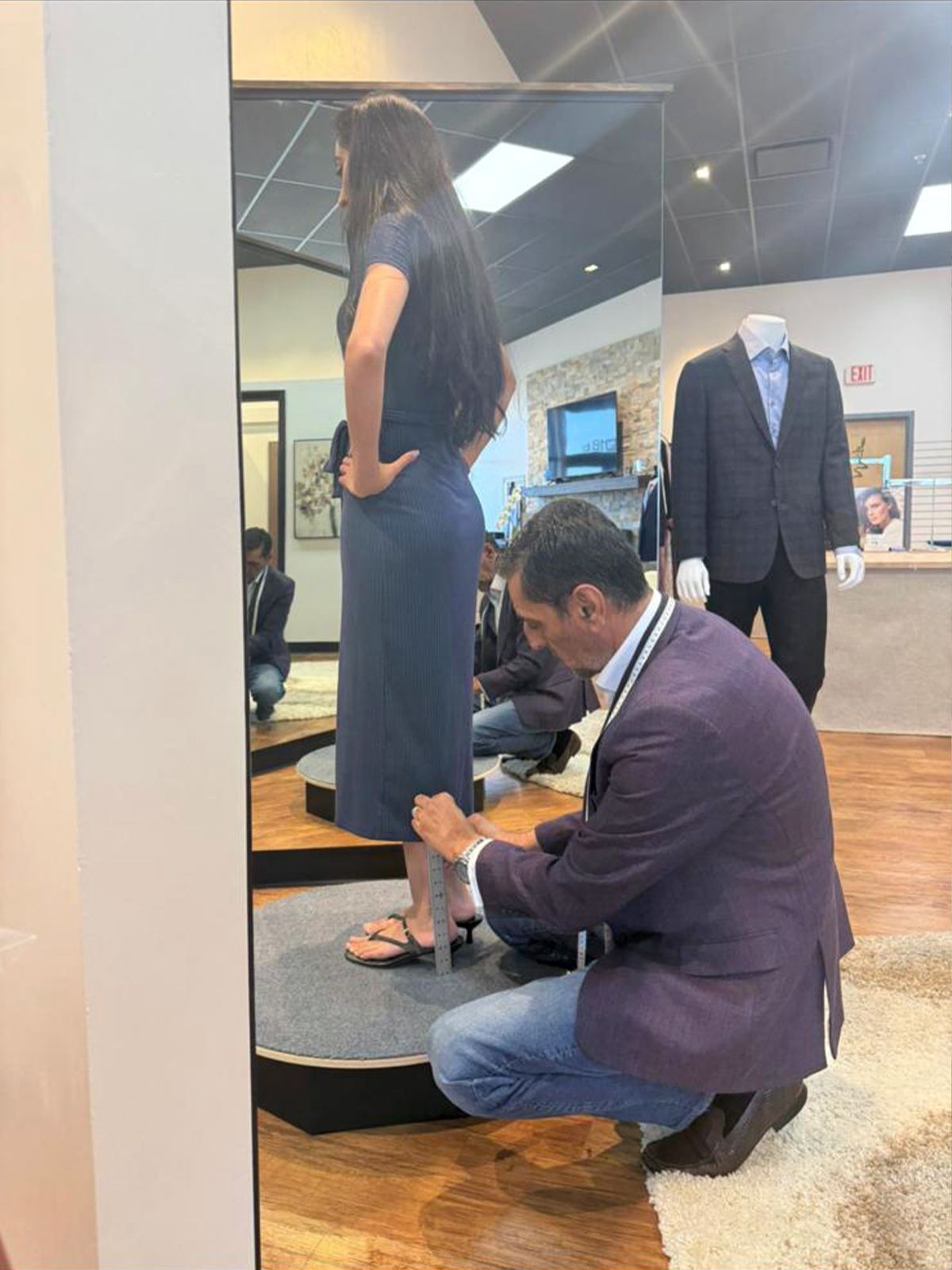 A tailor measures a woman's skirt length in front of a mirror, inside a clothing store.