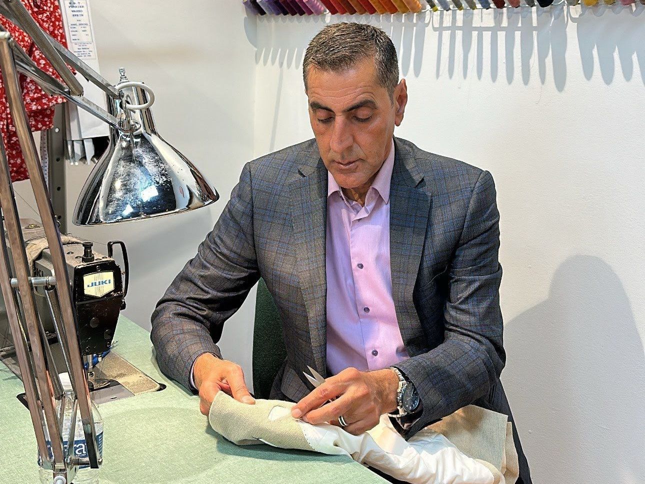 Man in blazer working on fabric at a sewing machine. Bright lamp and thread spools in the background.