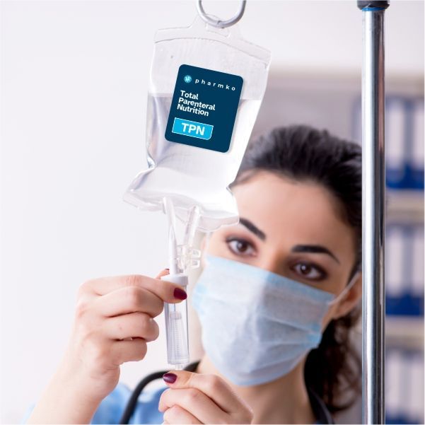 A healthcare professional wearing a surgical mask adjusts an IV bag labeled 'Pharmko Total Parenteral Nutrition (TPN)' in a clinical setting.

