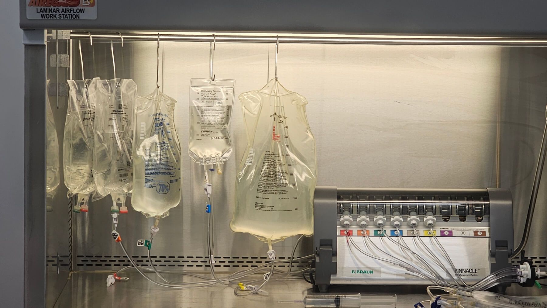 Sterile compounding workstation with multiple IV solution bags hanging inside a laminar airflow hood. Each bag is connected to tubing and labeled for various medications and nutrients. On the right side, a B. Braun Pinnacle compounder is visible, used for precision mixing of sterile IV formulations in a cleanroom environment.