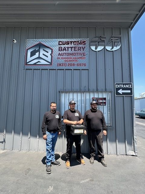Three men are standing in front of a building with a sign that says customs battery automotive.