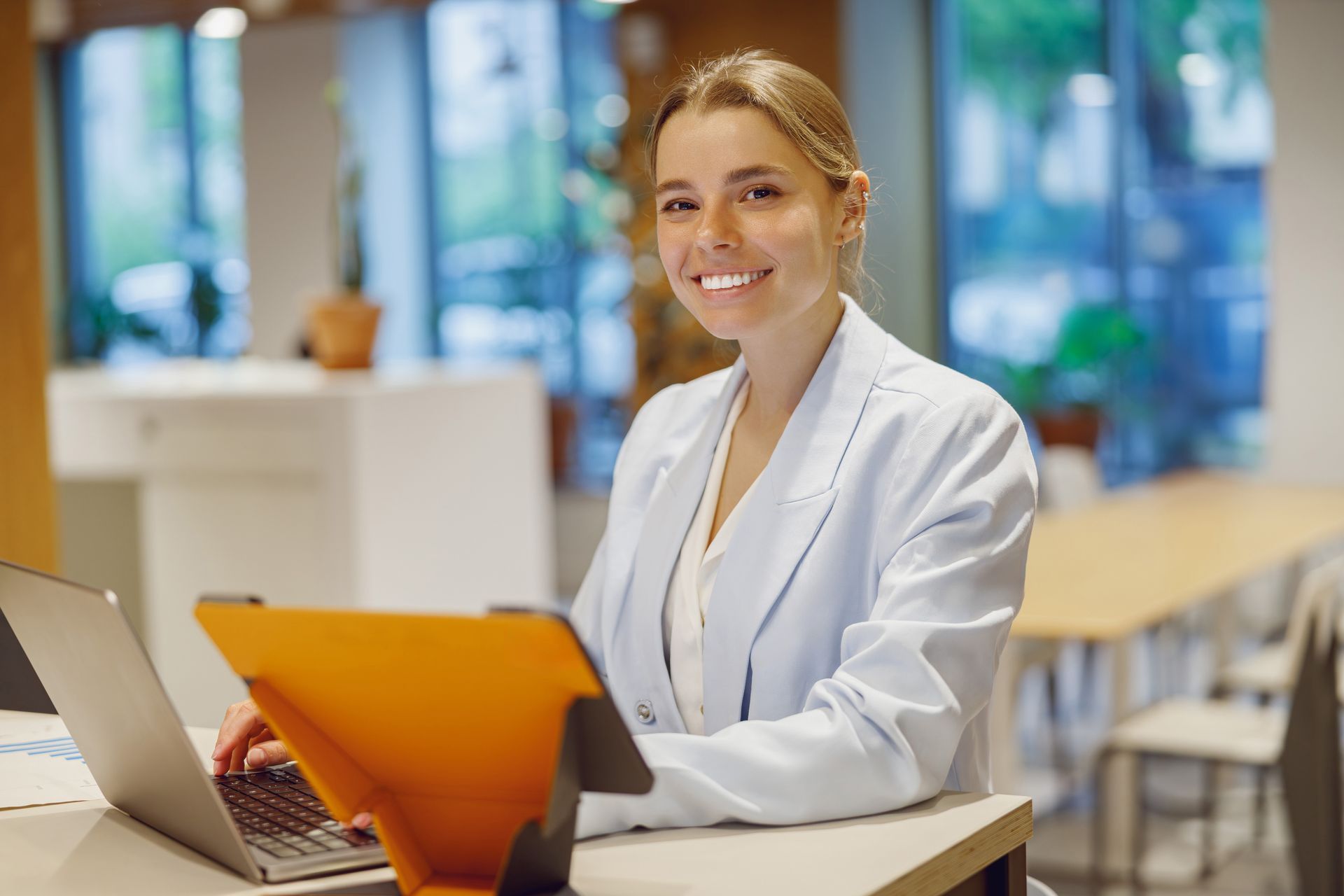 Woman smiling, working on laptop and tablet at a desk in an office.