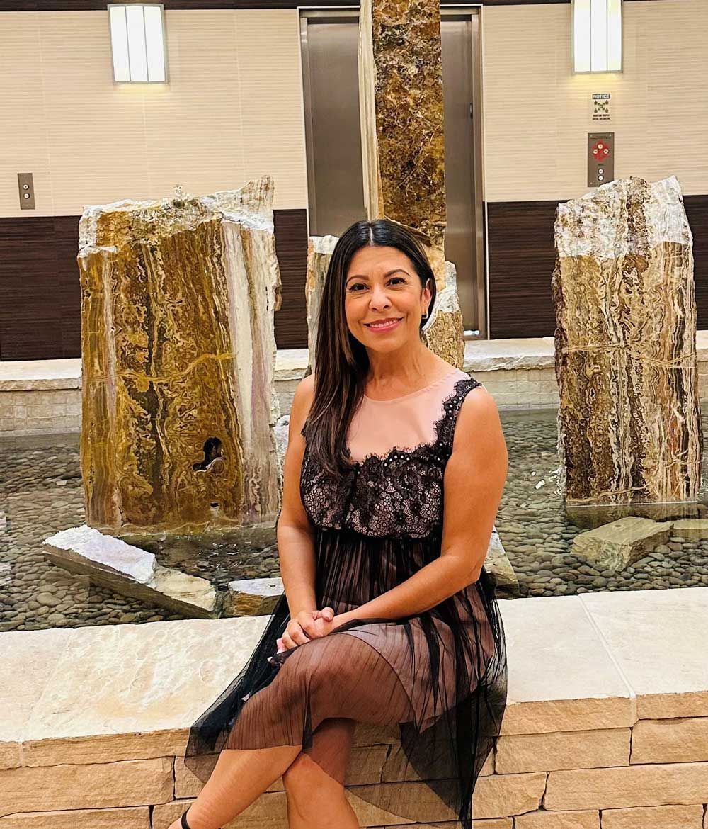 A woman in a black dress is sitting on a wall in front of a fountain.
