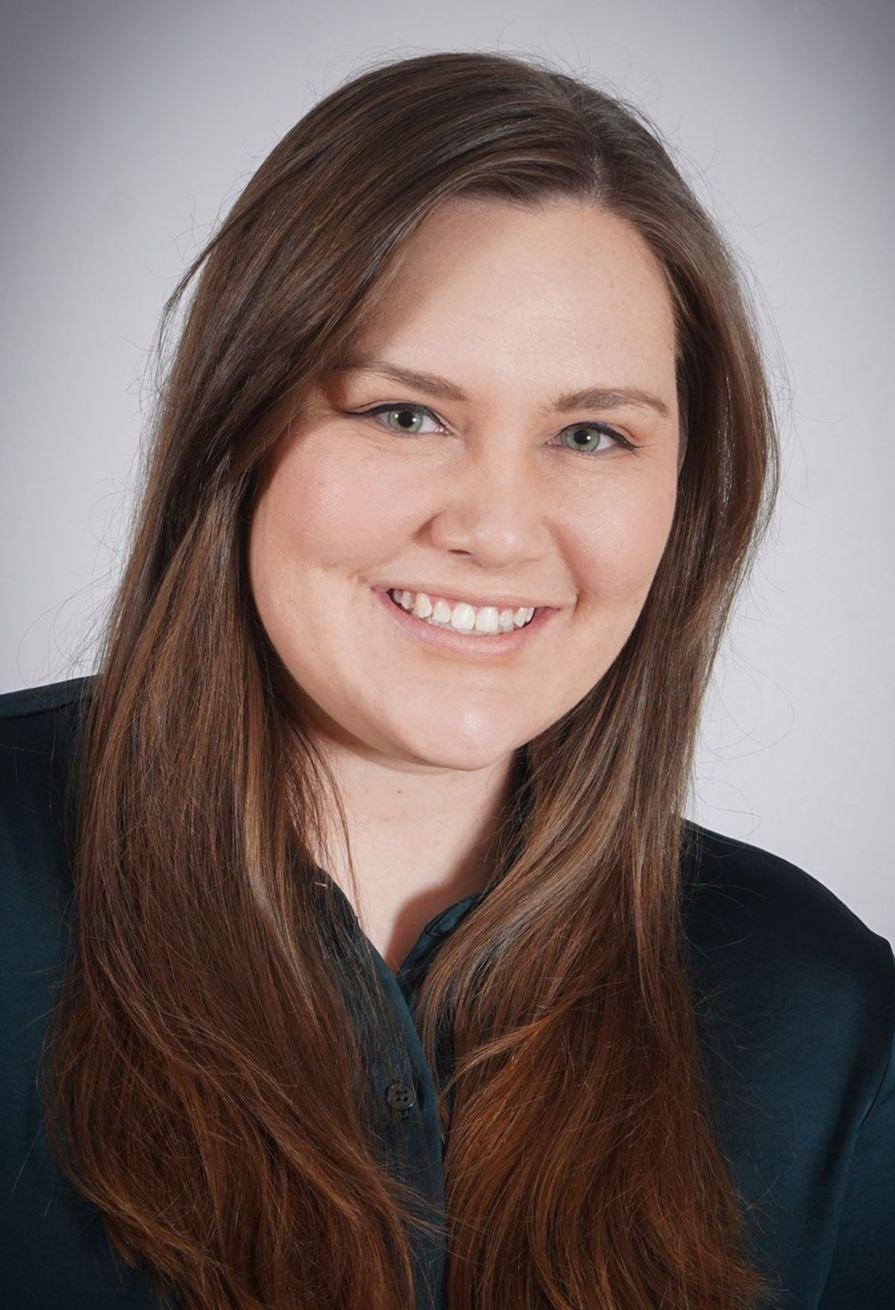 A woman with long brown hair is smiling and wearing a blue shirt.