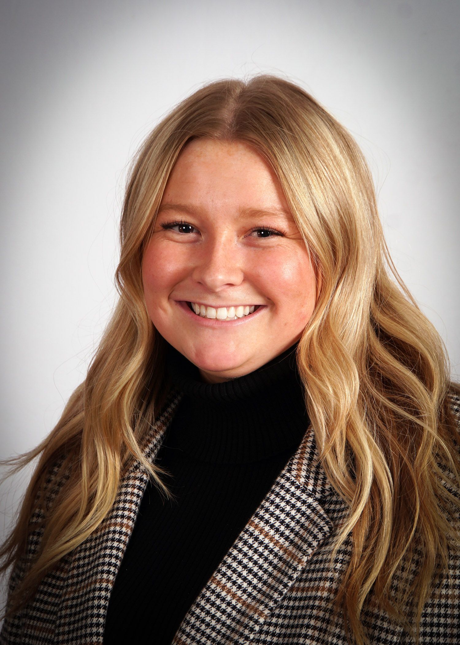 Woman with blonde hair smiling, wearing a black turtleneck and plaid blazer.