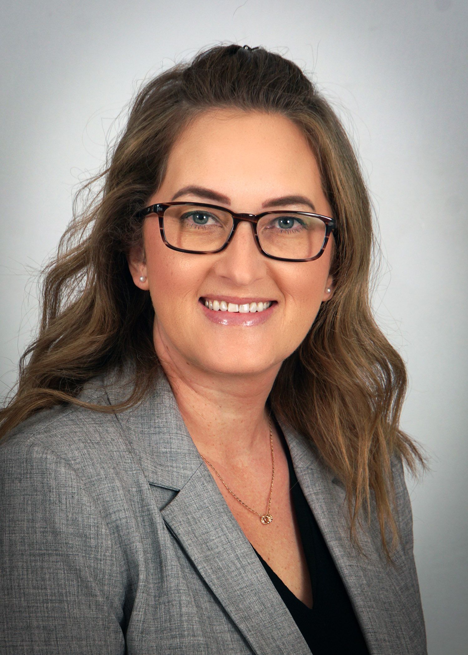 Woman in glasses and a gray blazer smiles, looking at the camera.
