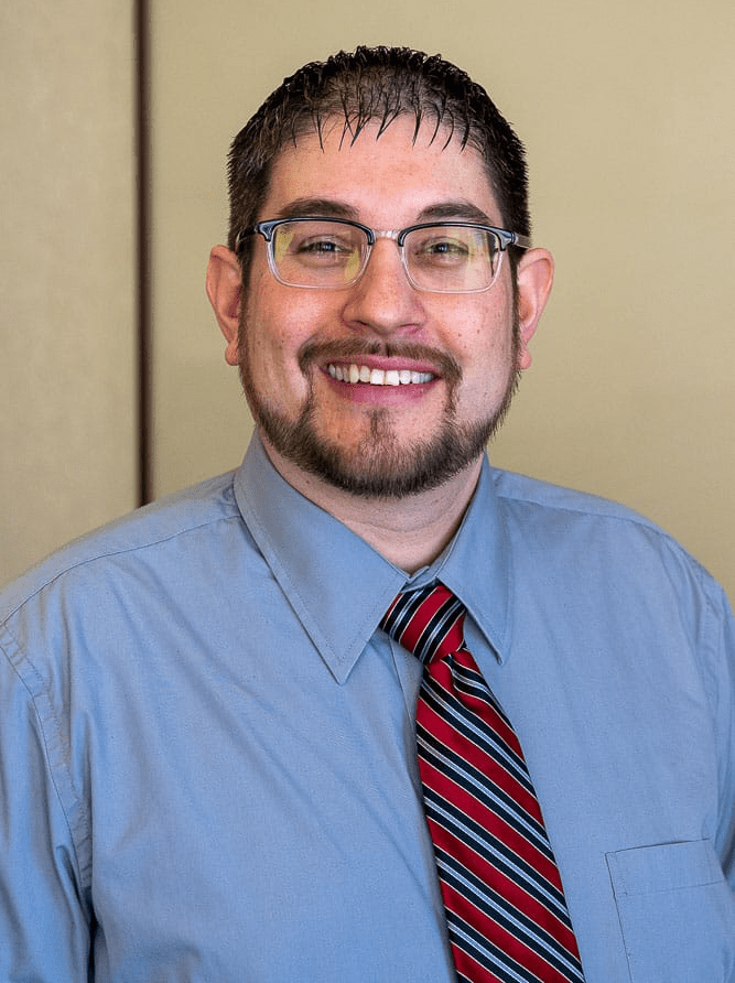 A man with glasses and a beard is wearing a blue shirt and red tie.
