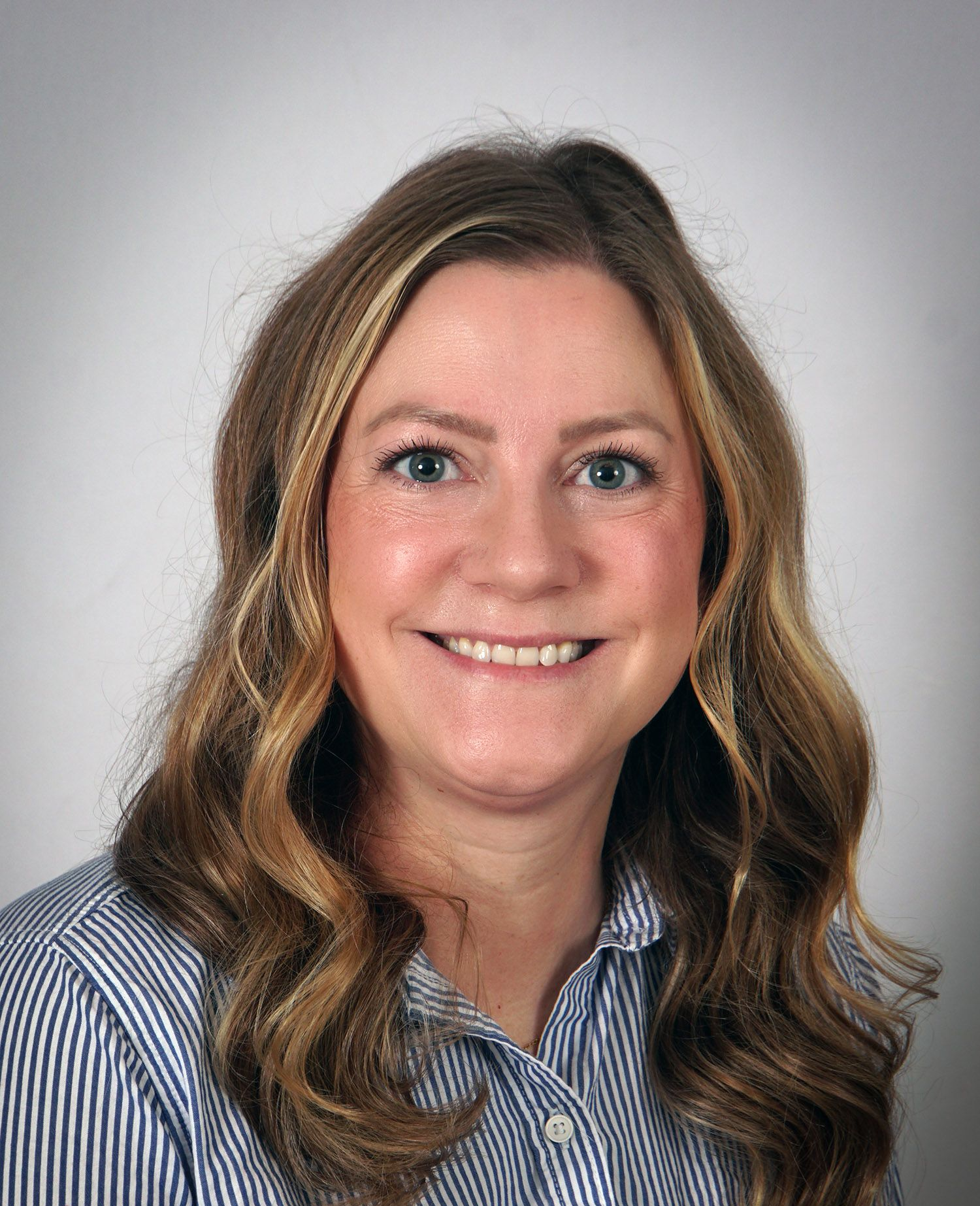 Woman with wavy blonde hair, smiling, wearing a striped blue and white button-down shirt.