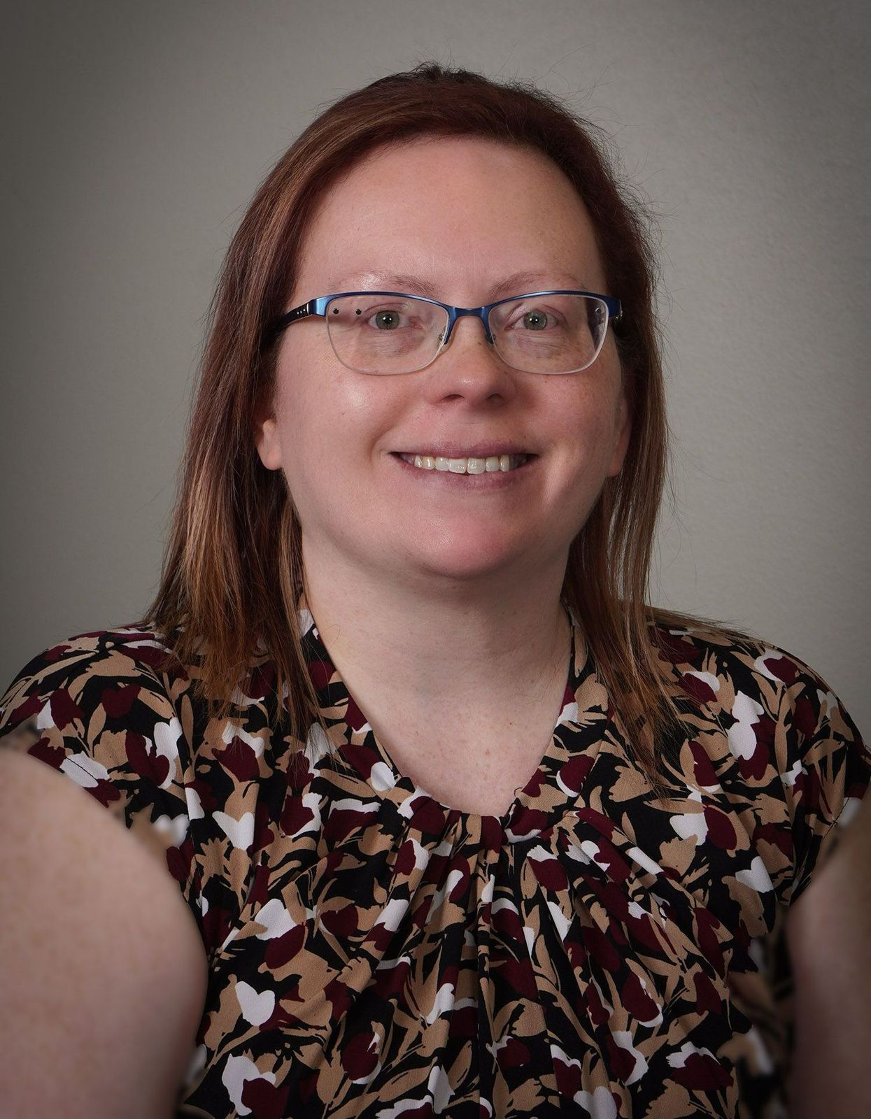 A woman wearing glasses and a floral shirt is smiling for the camera.