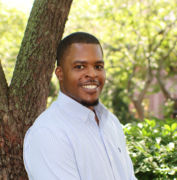 Smiling man in a light blue shirt leaning against a tree in a sunny garden