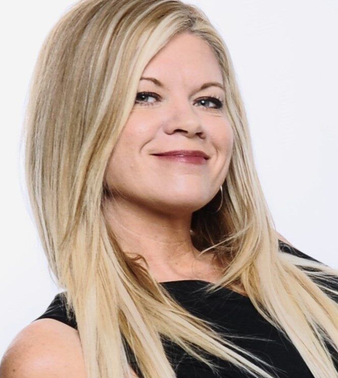 Woman with blonde hair smiles; wearing a black top, looking at camera.