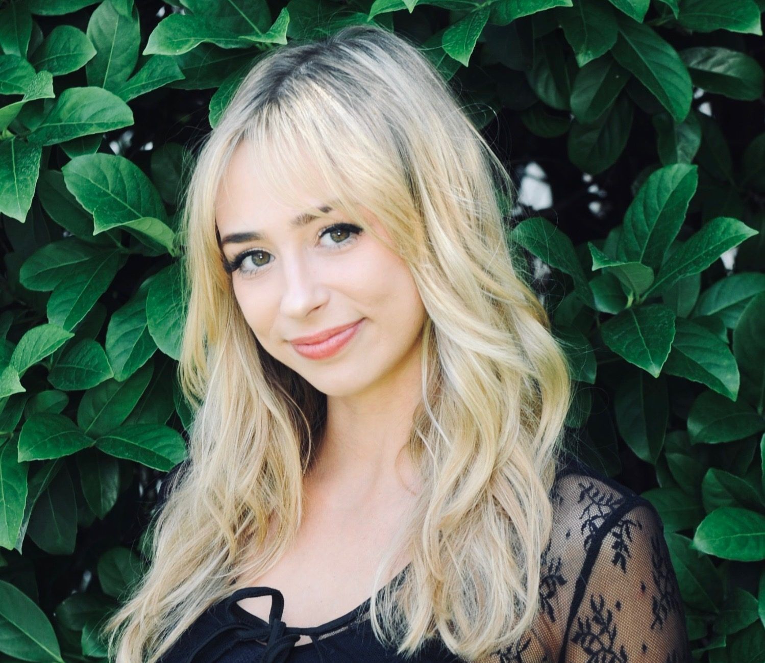 Blonde woman smiles in front of green foliage, wearing a black lace top.