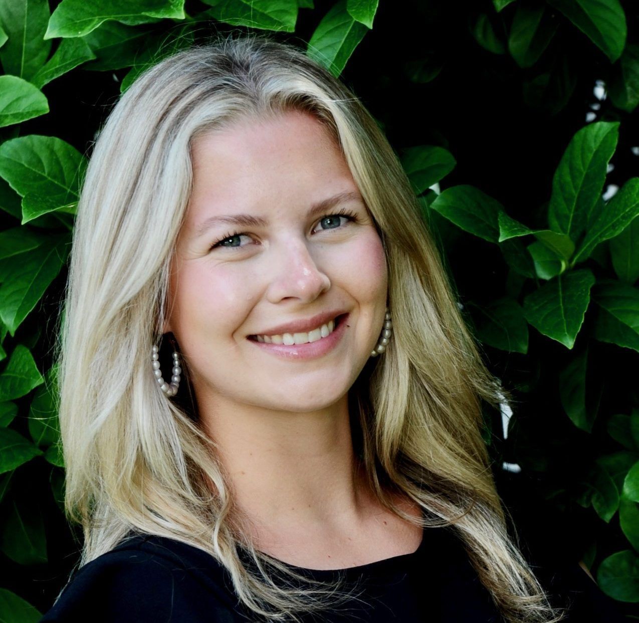 Blonde woman with a warm smile, wearing a black top, posed in front of green foliage.