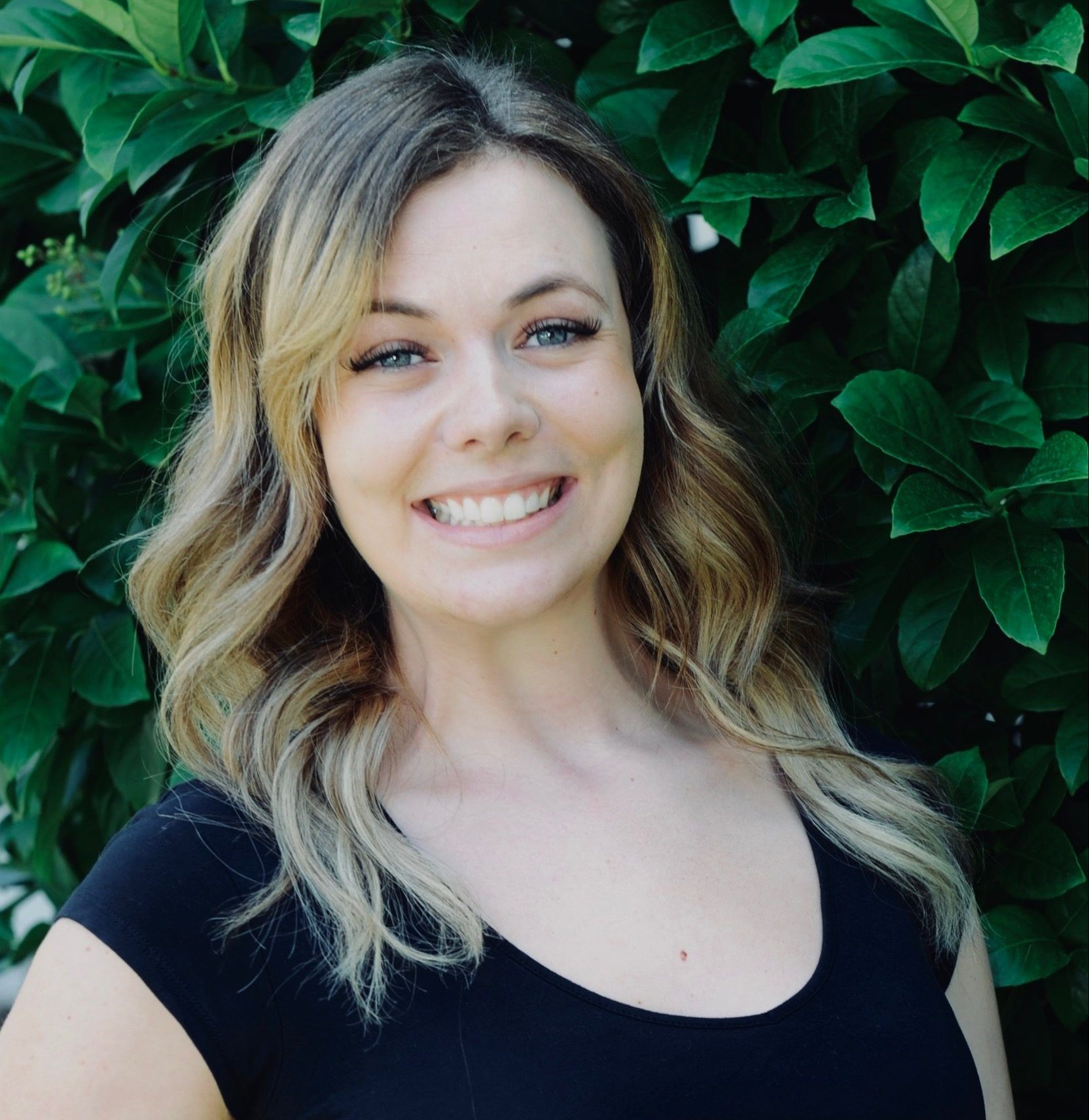 Woman with blonde hair smiling in front of green foliage. Wearing a black top.