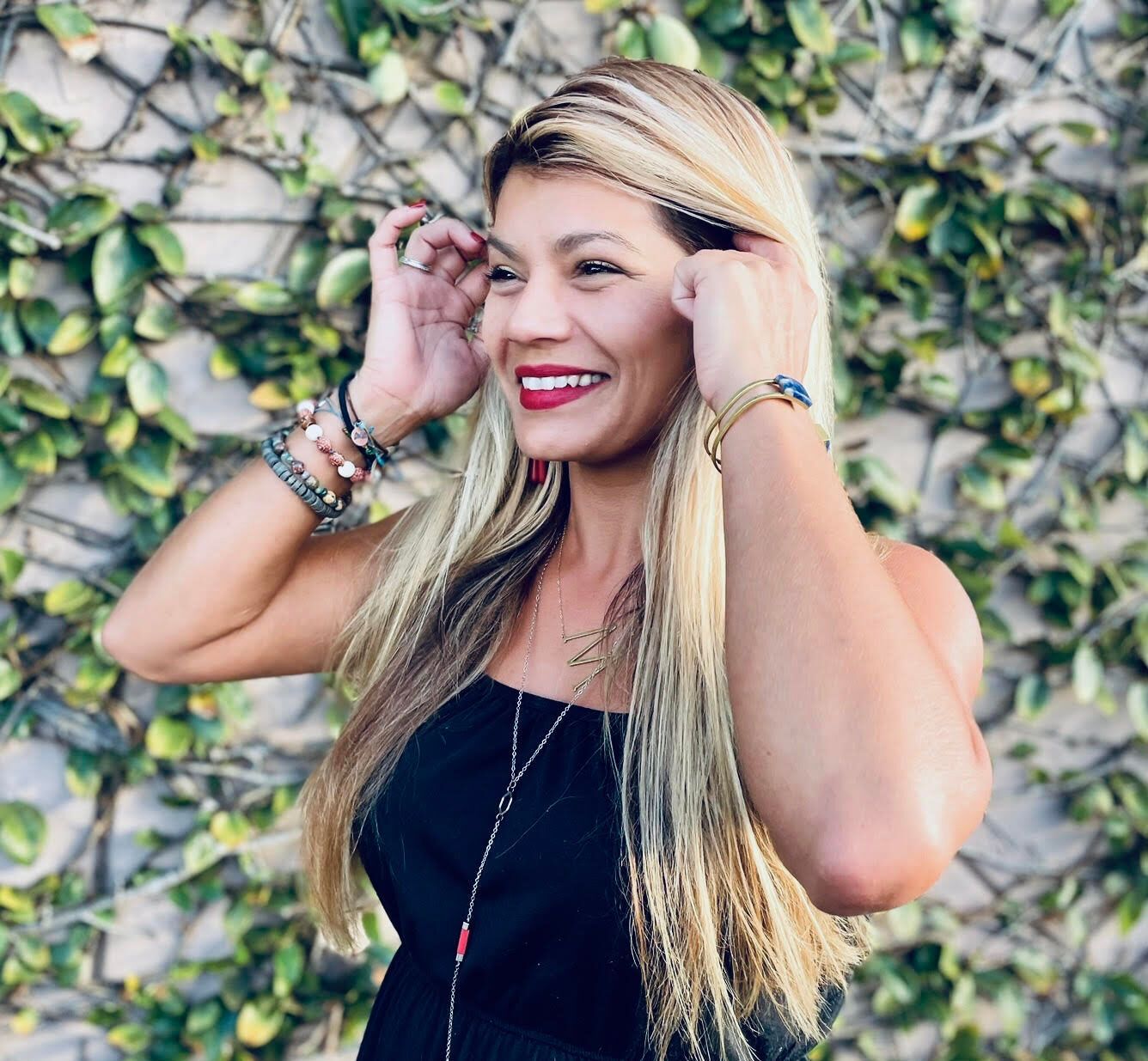 Woman with blonde hair, wearing jewelry and a black top, smiling, posing in front of a green wall.