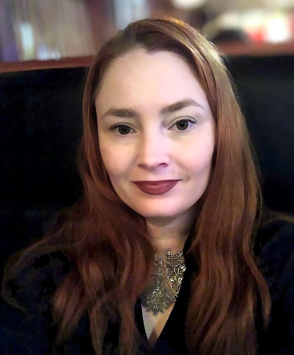 Woman with auburn hair, dark lipstick, and ornate silver necklace; seated, looking at the camera.