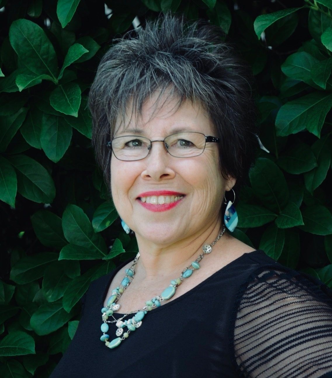 Smiling woman with glasses, short grey hair, blue necklace and earrings, in front of green leaves.