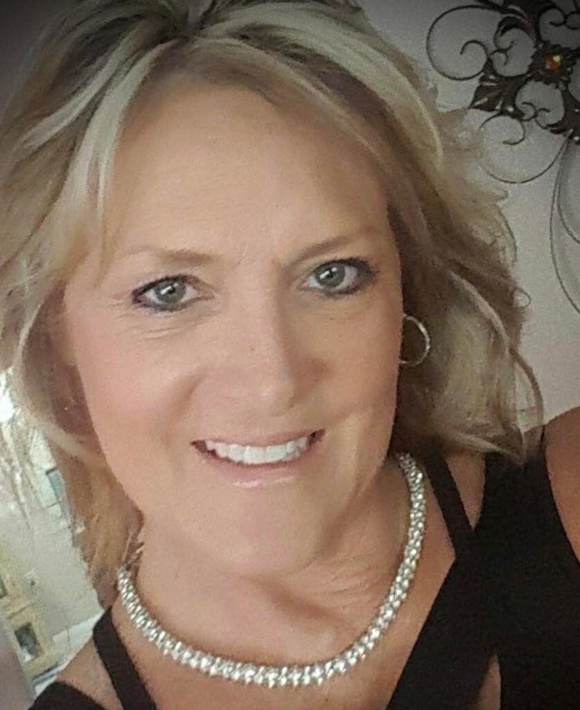 Blond woman with diamond necklace smiles, looking at the camera. Indoor setting.