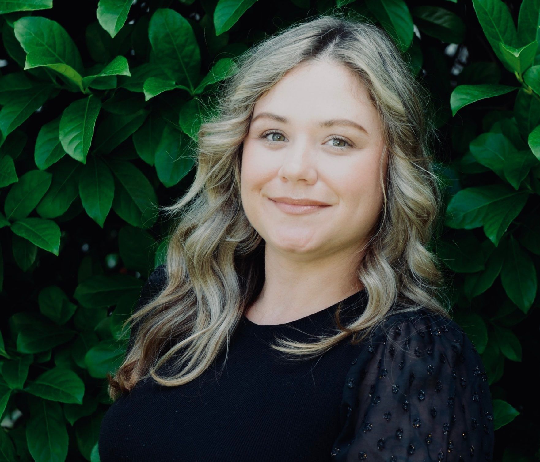 Woman with blonde wavy hair smiles in front of green leaves, wearing a black top.