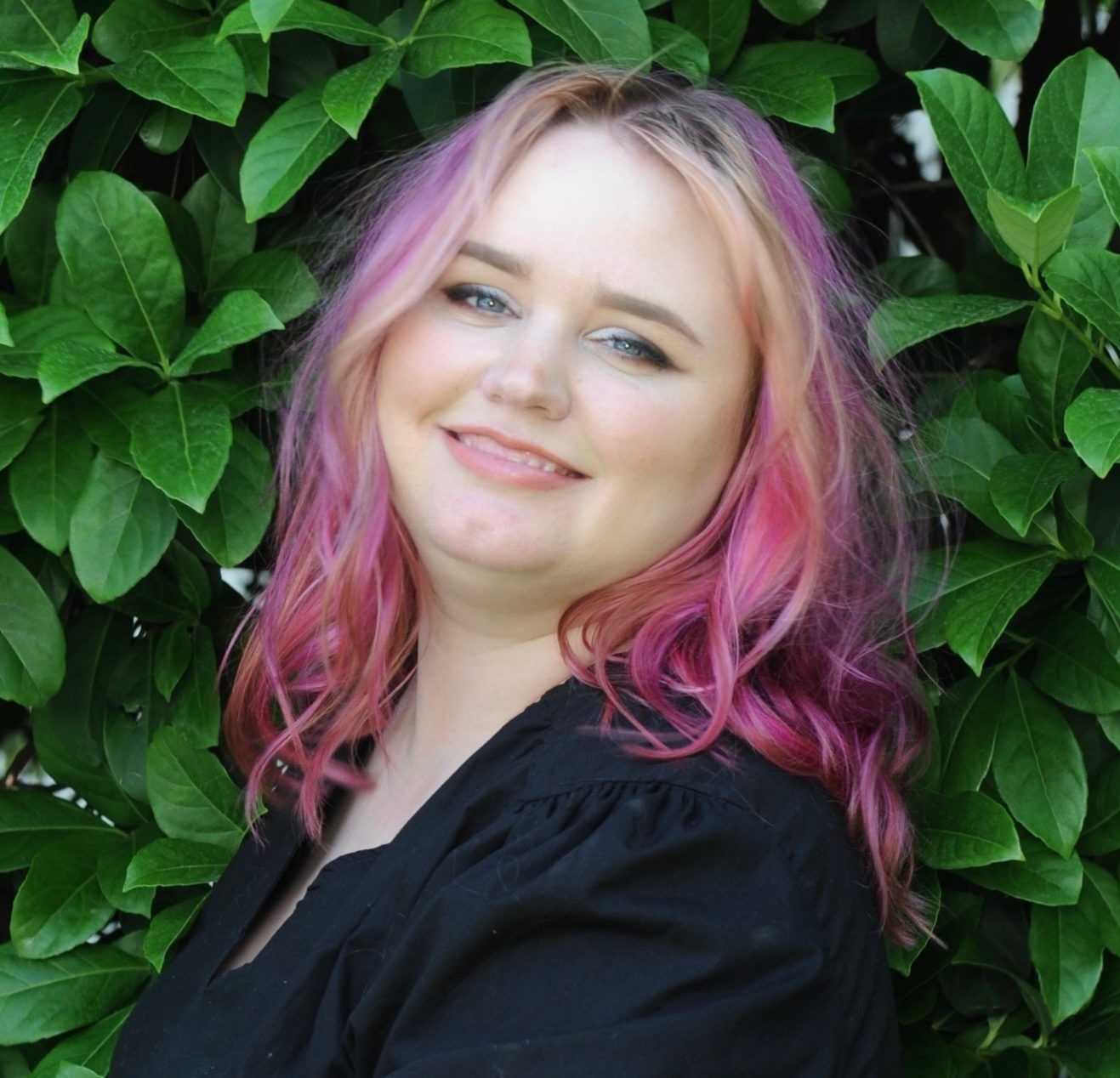Woman with pink hair smiles in front of green foliage.