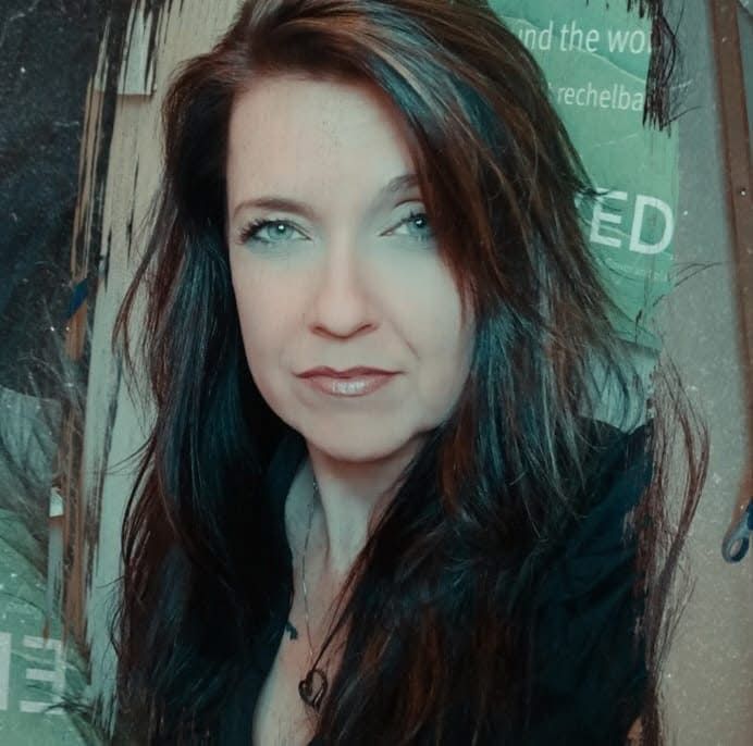 Woman with long brown hair and blue eyes, wearing a black top, in front of a wall of paper.